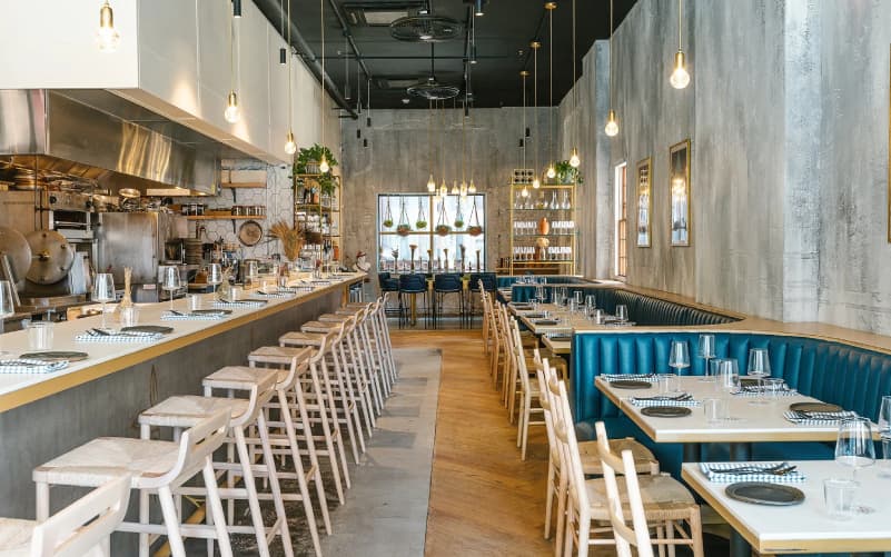 Interior of Krasi Greek wine bar in Back Bay, Boston, with white bar stools along a counter, teal banquette seating, and set tables.