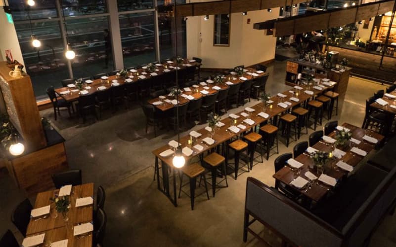 Interior of Paqu restaurant in Cambridge, with rows of dark wooden tables and stools, large windows, and pendant lighting.