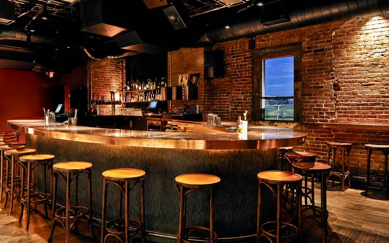 Interior of Brick & Mortar cocktail bar in Cambridge, with exposed brick walls, a curved bar lined with orange-topped stools, and dim lighting.