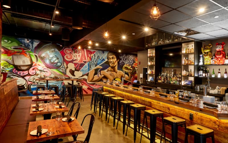 Interior of Shojo in Boston's Chinatown, with a colorful graffiti mural, a long wooden bar lined with stools, and wooden tables.