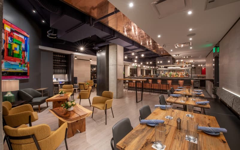 Interior of Alcove restaurant in Boston's West End, with wooden dining tables, mustard-toned upholstered chairs, a bar in the background, and colorful abstract art.