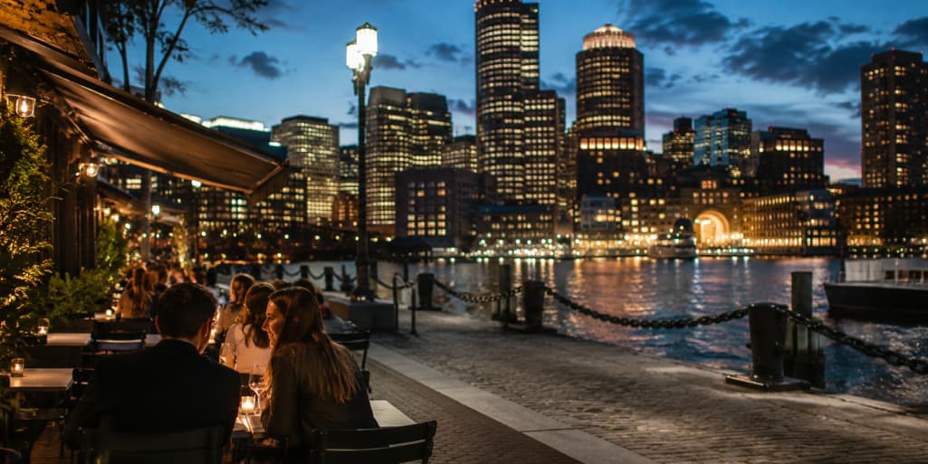 Couple dining on a waterfront patio at night with the illuminated Boston skyline across the harbor, representing a romantic first-date setting in the Seaport.