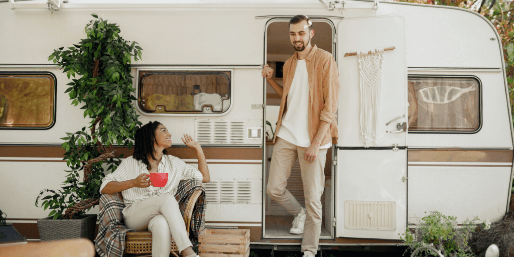 A woman sitting down drinking a hot beverage and a man walking toward her from inside the camper or tiny house.