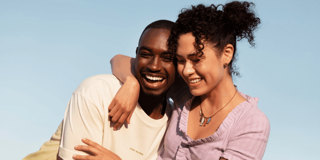 Don't let your insecurities get in the way. A man and woman embrace each other and laugh with blue skies in the background.