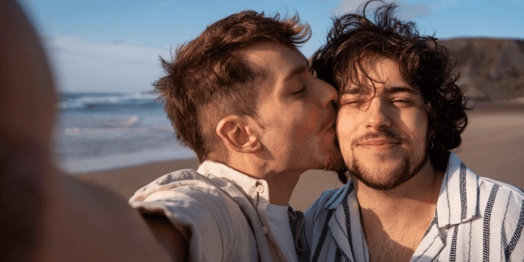 How men can overcome commitment issues. A man kisses his boyfriend as they take a selfie of themselves at the beach.